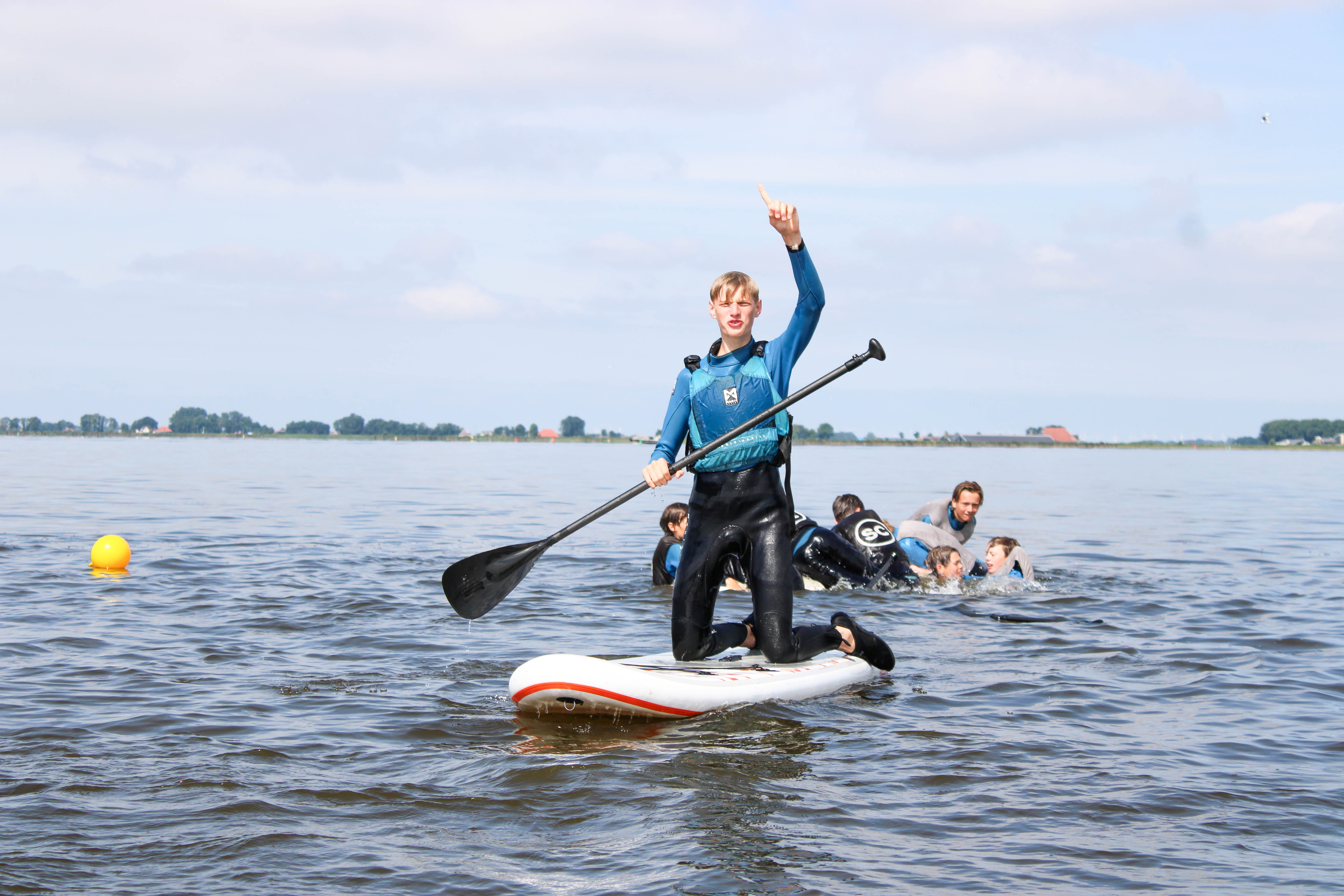Een tiener in een wetsuit en reddingsvest knielt op een supboard op het water en wijst omhoog, met op de achtergrond andere kinderen die in het water spelen