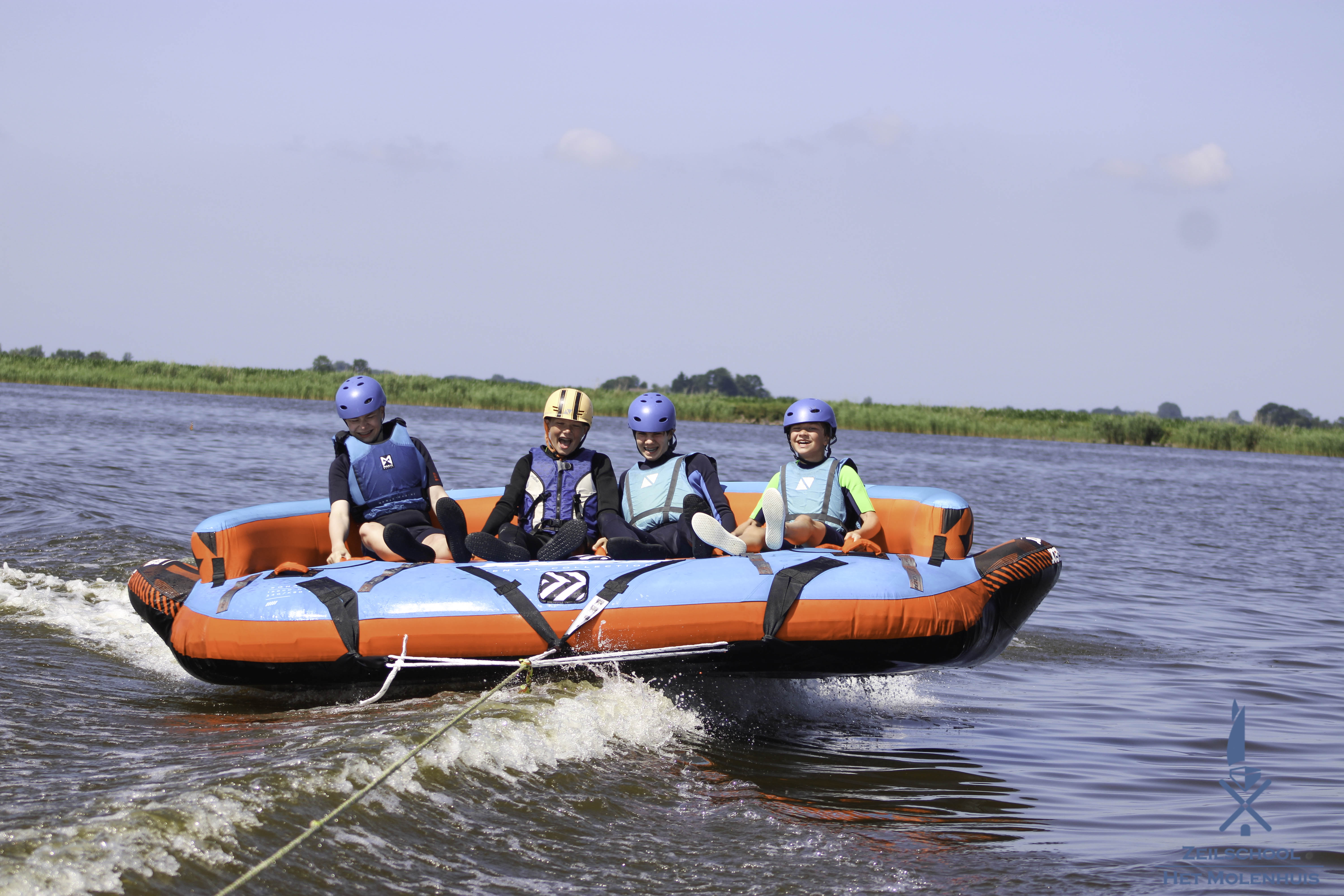 Vier blije kinderen met helmen en zwemvesten op een blauw-rode tube op het water