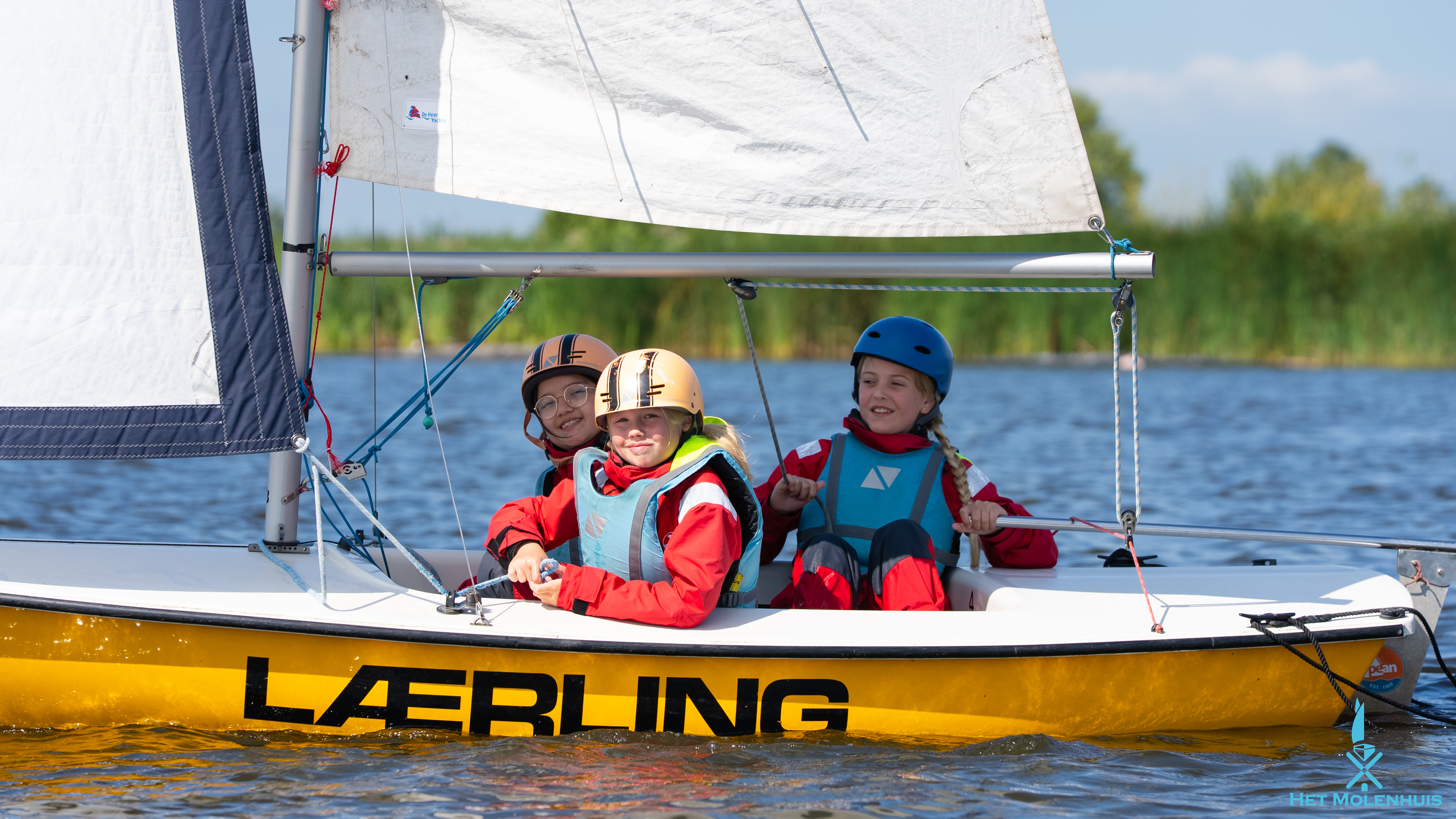 Drie meisjes met helmen, rode zeiljassen en zwemvesten in een gele LÆRLING-zeilboot op het water